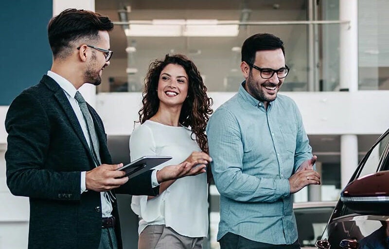 A saleman smiling while greeting couple looking at car.